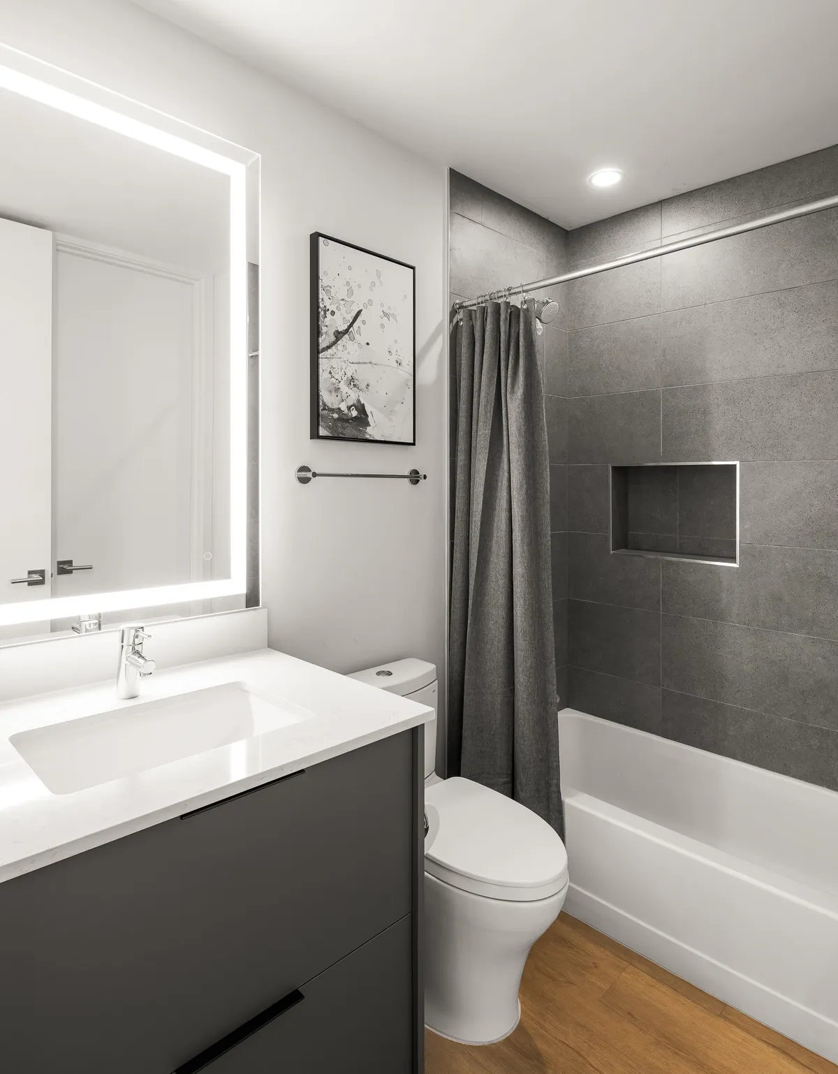 A contemporary bathroom with a bathtub behind a grey shower curtain, framed artwork, and a backlit mirror over a modern sink. The ambiance is serene and inviting.