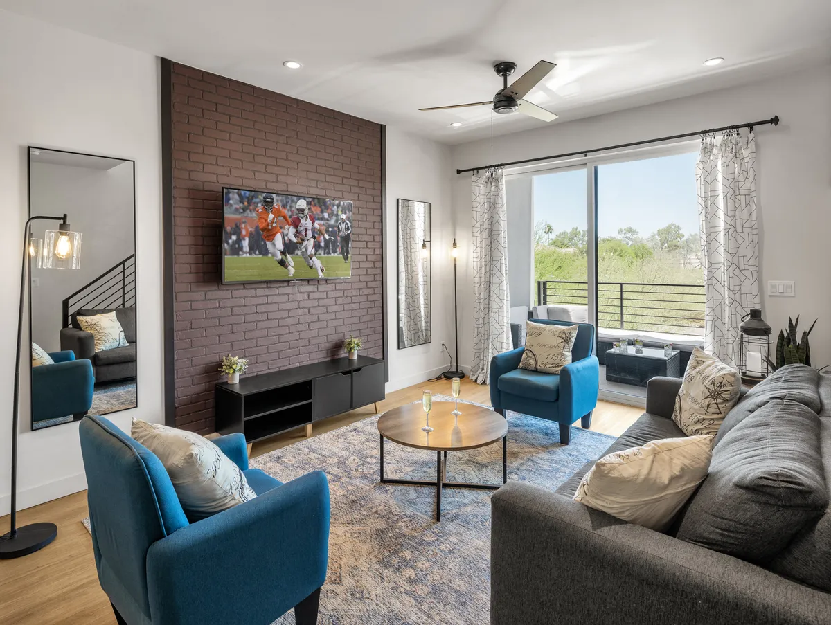A comfortable living area featuring plush blue armchairs, a sofa, a coffee table, a wall-mounted television, and a large window providing natural light and a view. The setup is ideal for relaxing, entertainment, or catching up after a day out.