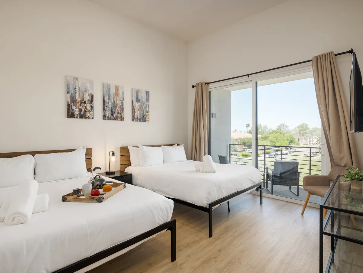 A bright bedroom with two beds, decorative wall art, and a direct view to a private balcony, suggesting access to outdoor space and plenty of natural light. The setting feels inviting, ideal for quiet mornings or wind-down evenings.