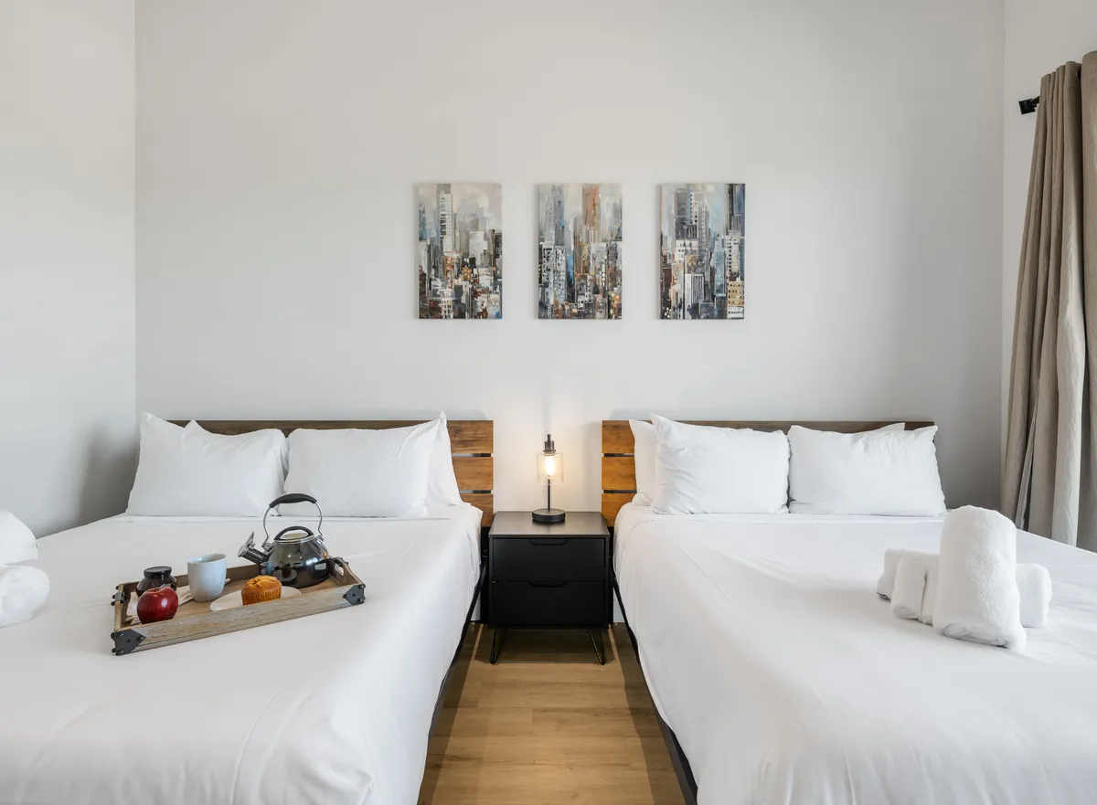A bright and airy bedroom featuring two well-made beds with decorative artwork above, a nightstand with a lamp, and a breakfast tray. The décor is comfortable yet refined, emphasizing cleanliness and attention to detail.
