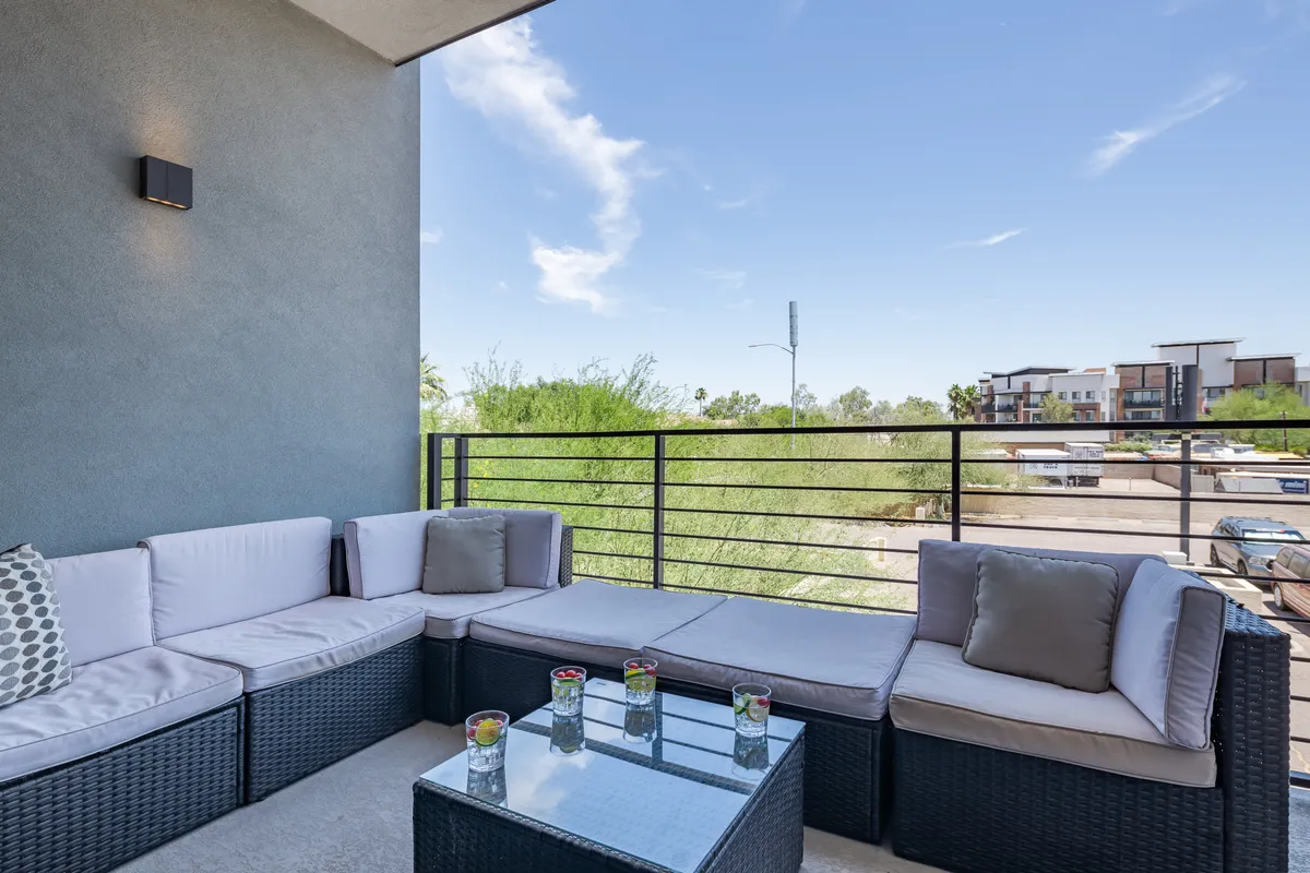 A serene exterior patio with a glass table and comfortable outdoor seating, framed by greenery in the background. The environment is perfect for enjoying a peaceful morning coffee, casual meetings, or winding down in the evening. The ambiance is quiet and welcoming, ideal for individual relaxation or informal colleague interactions.