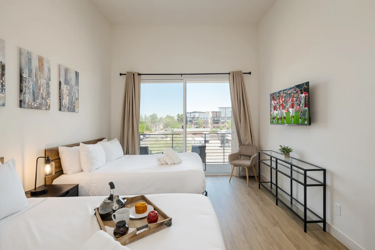 This interior image captures a spacious bedroom with two beds, a large window allowing natural light, a TV, and decorative artwork. The setting feels contemporary and comfortable, ideal for relaxation or entertainment.