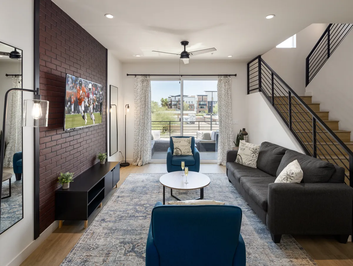 The image showcases a stylish and comfortable living room with a prominent brick accent wall, a large television, a contemporary sofa facing a sleek coffee table, and expansive windows that bathe the space in natural light. The inviting setup encourages lounging, socializing, and enjoying scenic views, while modern décor blends warmth and sophistication.