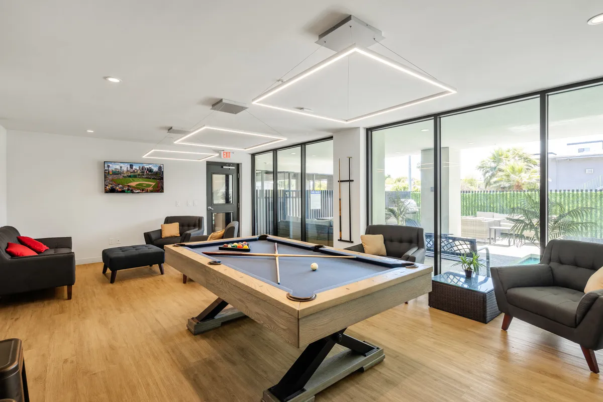 A spacious clubhouse interior featuring a pool table in the center, comfortable sofa seating, and large windows that let in ample natural light. The relaxed layout encourages socializing and leisure activities, making it ideal for gatherings and friendly competition.